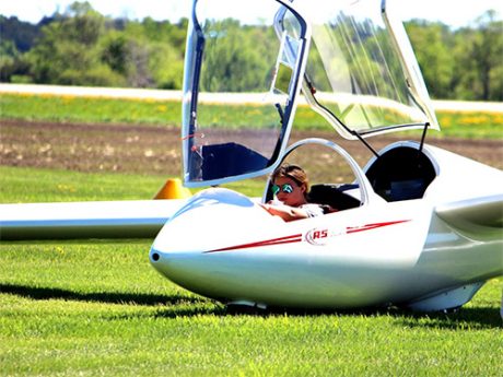 A pilot in the open cockpit of a glider on a grass field